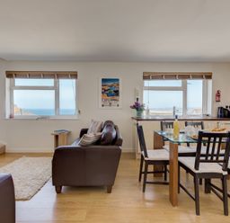 A living room and kitchen with a brown sofa wooden chairs and a glass dining table at Atlantic Blue in Carbis Bay
