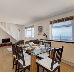 A dining area with a glass table set for breakfast and a living room with sofas and a television near a staircase at Atlantic Blue in Carbis Bay