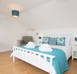 A bedroom with a white bed and turquoise bedding with pillows and white towels on the bed with a striped armchair and white bedside tables at Atlantic Blue in Carbis Bay