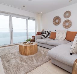 A living room with a grey sofa and cushions a wooden round coffee table a beige rug and glass doors overlooking the sea at Atlantic Watch 8 in Carbis Bay
