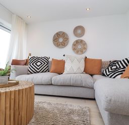 A living room with a gray sectional sofa with patterned and solid cushions a round wooden coffee table and woven wall decor at Atlantic Watch 8 in Carbis Bay