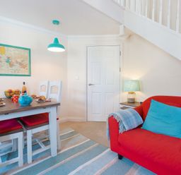 A dining area with a wooden table and white chairs with red cushions and a red sofa with blue cushions under a staircase at Brinyloft in Carbis Bay