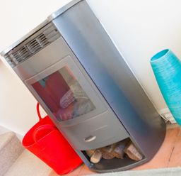 A metal wood stove with logs underneath a red basket and a blue vase standing on a tiled floor at Brinyloft in Carbis Bay