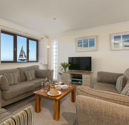 A living room with two sofas a TV on a stand a wooden coffee table with food and drinks and a window with a sailboat decoration at Cornerways in Carbis Bay