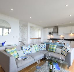 A living room with a grey sectional sofa and kitchen with white cabinets in the background at Halcyon Cove in Carbis Bay