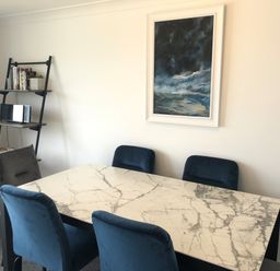 A dining room with a marble table and blue chairs a ladder shelf with books and a framed painting on the wall at Ocean Cove in Carbis Bay