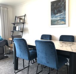 A dining room with a marble table and blue chairs a gray armchair with blue cushions a black leaning shelf with books and a stereo and a landscape painting on the wall at Ocean Cove in Carbis Bay
