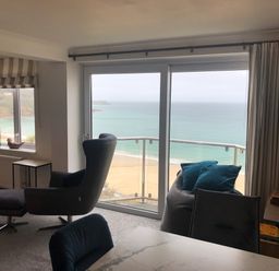 A living room with an armchair and ottoman near large windows overlooking a beach at Ocean Cove in Carbis Bay