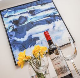 A bottle of red wine with two wine glasses and yellow flowers in a vase in front of a blue abstract painting on a wall