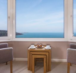 Two chairs and nested tables with cups and a plate of pastries by large windows overlooking the sea at Seathrift in Carbis Bay