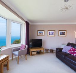 A living room with a large window overlooking the ocean a leather sofa with cushions a TV on a wooden stand a chair and nested wooden tables at Seathrift in Carbis Bay