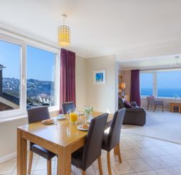 A dining table set with croissants and juice near a window with a view of houses and a living room with chairs and a sea view at Seathrift in Carbis Bay