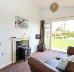 A living room with a brown leather sofa a small black fireplace a wall-mounted TV and large sliding glass doors at Chalet 150 in Winterton-On-Sea