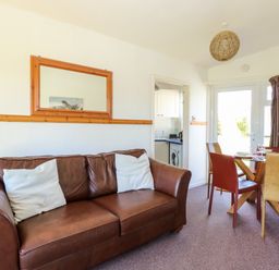 A living room with a brown leather sofa two white cushions a wooden framed mirror and a dining table with four chairs and flowers at Chalet 150 in Winterton-On-Sea
