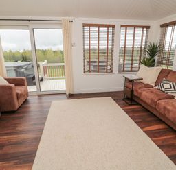 A living room with brown sofas and large windows showing greenery outside at Woodland View Lodge Swarland near Felton