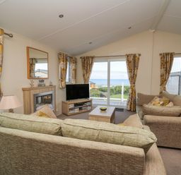 A living room with two sofas a coffee table a TV on a stand a fireplace and yellow floral curtains with a view outside at The Potting Shed in New Quay