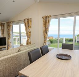 A dining table with chairs and a couch facing a television near windows with curtains showing a view of the sea at The Potting Shed in New Quay