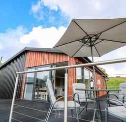 An outdoor patio with cushioned chairs a table with a wine bottle and glass and a large umbrella attached to a modern house at Llyn Cae Ty Nant Fishery Holiday Let Bwlchllan near Llangeitho