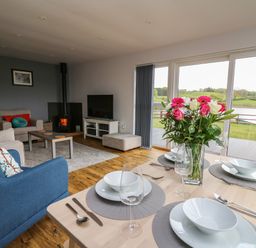 A dining table set with plates bowls glasses and cutlery next to a living room with armchairs a coffee table a wood stove and a television at Llyn Cae Ty Nant Fishery Holiday Let in Bwlchllan near Llangeitho
