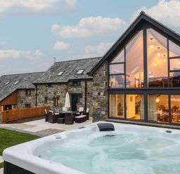 A stone house with large windows next to a patio with outdoor furniture and a hot tub at Merddwr in Cerrigydrudion