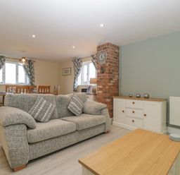 A living room with a gray sofa, wooden coffee table, white sideboard, brick chimney, dining table with chairs, and windows with patterned curtains at Bramley Cottage in Henley near Langport