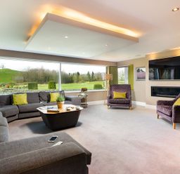 A living room with grey sofas, purple armchairs, a coffee table, a wall-mounted television, and large windows at Carus House at Carus Green Golf Club Kendal