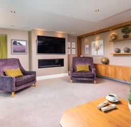 A living room with two purple armchairs facing a wall-mounted TV and a modern fireplace at Carus House at Carus Green Golf Club in Kendal