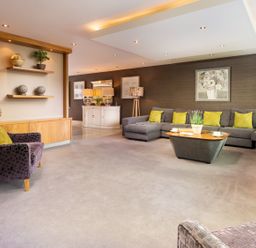 A living room with a large gray sectional sofa with yellow pillows a purple armchair two wooden shelves with plants and decorative objects a wooden coffee table with a plant and cups a white cabinet with lamps and framed pictures on the walls at Carus House at Carus Green Golf Club in Kendal
