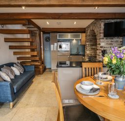 A dining area with a round wooden table set with dishes and a vase of flowers next to a kitchen with wooden beams and a blue sofa at Swallows Nest in Whitby