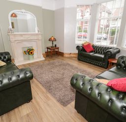 A living room with three dark green leather sofas a beige rug wooden flooring a white fireplace with a mirror and a bouquet of sunflowers on it a side table with a lamp and large windows with white blinds at 23 Carmen Sylva Road in Llandudno