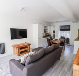 An open plan living room and kitchen with a gray sofa facing a wall mounted TV wooden dining table with four chairs and white kitchen cabinets at 8 Waterhead Terrace Ambleside