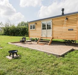 A wooden shepherds hut with steps and outdoor chairs on a gravel area with grass and a fire pit at Waylands in Uffington