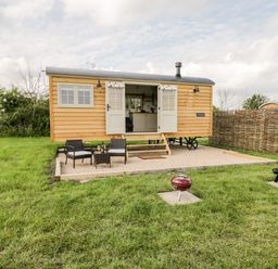 A small wooden cabin with open double doors on a gravel patio with two chairs a table and a red grill on grass at Waylands in Uffington