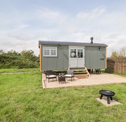 A small grey corrugated metal cabin with double doors and windows on a gravel patio with two chairs and a table on grass at Ridgeway in Uffington