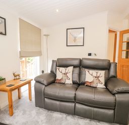 A living room with a leather sofa with deer cushions a wooden side table with a plant and decoration and framed deer pictures on the wall at Robin Lodge in Cartmel