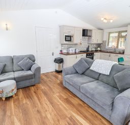 A living room with two grey sofas and a small ottoman in front of a kitchen with cabinets and a dining table at Grizedale Lodge in Langdale 18 White Cross Bay near Windermere