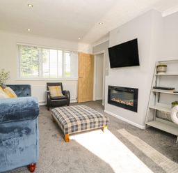 A living room with a blue sofa yellow cushions a plaid ottoman a wall-mounted TV an electric fireplace and a white shelving unit at 26 Hycemoor Way Bootle Near Millom