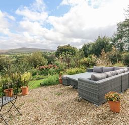 An outdoor seating area with a gray wicker sofa and tabe a black metal chair and potted plants on gravel at Nattaden @ Fourwinds in Meldon near Okehampton