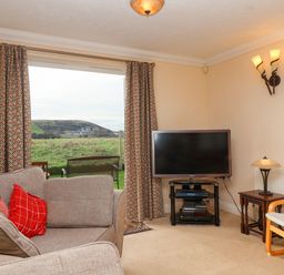 A living room with a sofa, television on a stand, armchair, side table, lamp, and large window with curtains at West Bay Holiday Home in West Bay