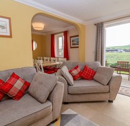 A living room with gray sofas and red cushions next to a dining area with white chairs and a window at West Bay Holiday Home in West Bay
