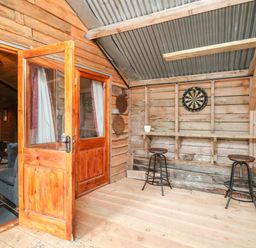 A rustic wooden room with a dartboard a wooden bar with two stools and an open door leading to a living room at Y Caban Pontfadog near Llangollen