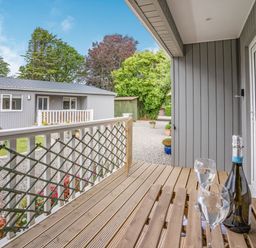 A wooden porch with a table holding a bottle and two glasses outside a gray cabin at Caldey Lodge (Bier Wood Lodges) in Manorbier