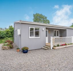 A small grey lodge with a front porch, flower boxes, and gravel driveway at Caldey Lodge Bier Wood Lodges in Manorbier