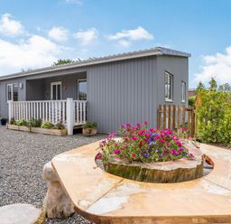 A grey lodge with white porch railing surrounded by gravel and a wooden bench with flowers in the center at Ramsey Lodge Bier Wood Lodges in Manorbier