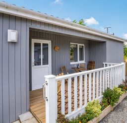 A porch with wooden chairs and table outside a grey house with white door and window at Ramsey Lodge Bier Wood Lodges in Manorbier