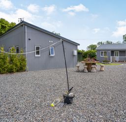 A yard with a badminton net and rackets between two gray buildings with a stone table and stools at Ramsey Lodge Bier Wood Lodges in Manorbier