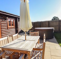 An outdoor patio with a wooden table and chairs and a closed umbrella next to a hot tub and a wooden fence at Holly Lodge in Greetham Rutland