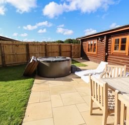 An outdoor patio area with wooden table and chairs white lounge chairs hot tub and wooden fence next to a log cabin at Pine Lodge in Greetham Rutland