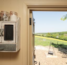 A wall-mounted cabinet with glass jars inside a room with an open door overlooking a green field and a tripod barbecue at The Shepherds Hut at Marley in Marley near Barham Kent