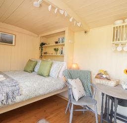 A small bedroom with a bed with green pillows a chair a small table with a basket and sunflowers and shelves with dishes and books at The Shepherds Hut at Marley